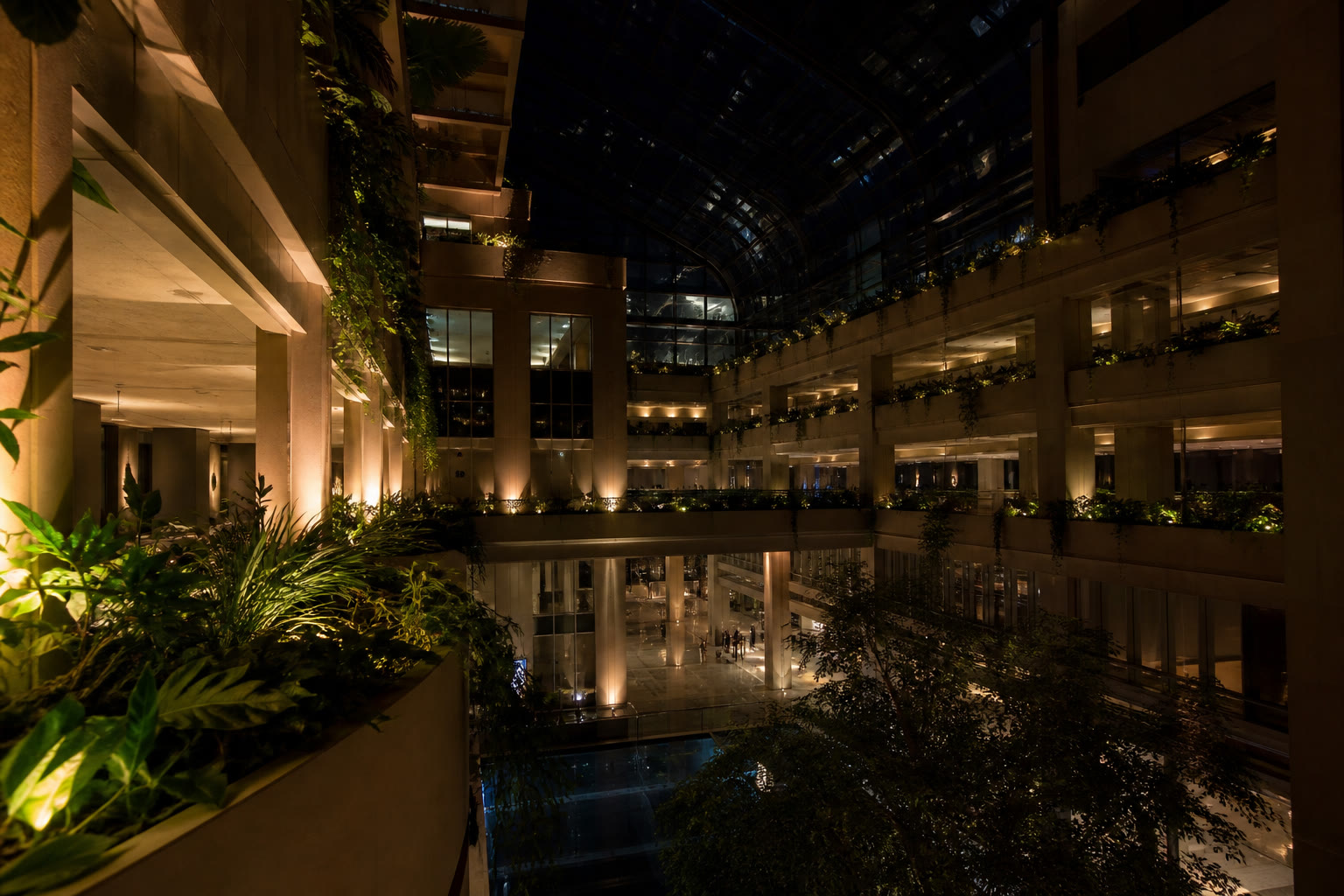A tall commercial atrium at night seen from a balcony looking across and down, planting on the levels and warm uplighting washing the walls.