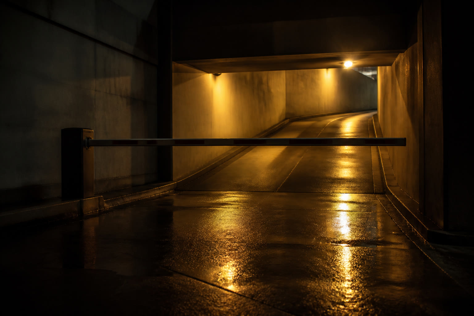 A car park exit ramp at night from a low angle, wet asphalt, a boom barrier lowered and warm sodium light spilling down the ramp behind it.