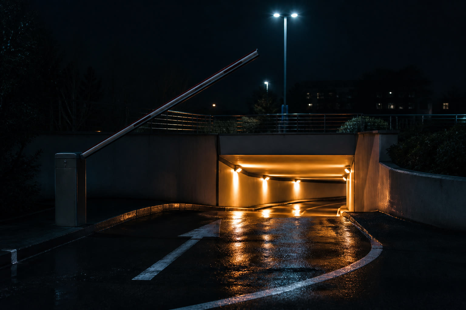 The entrance to an underground car park at night, a raised boom barrier over wet asphalt reflecting the overhead lights of the lit ramp beyond.