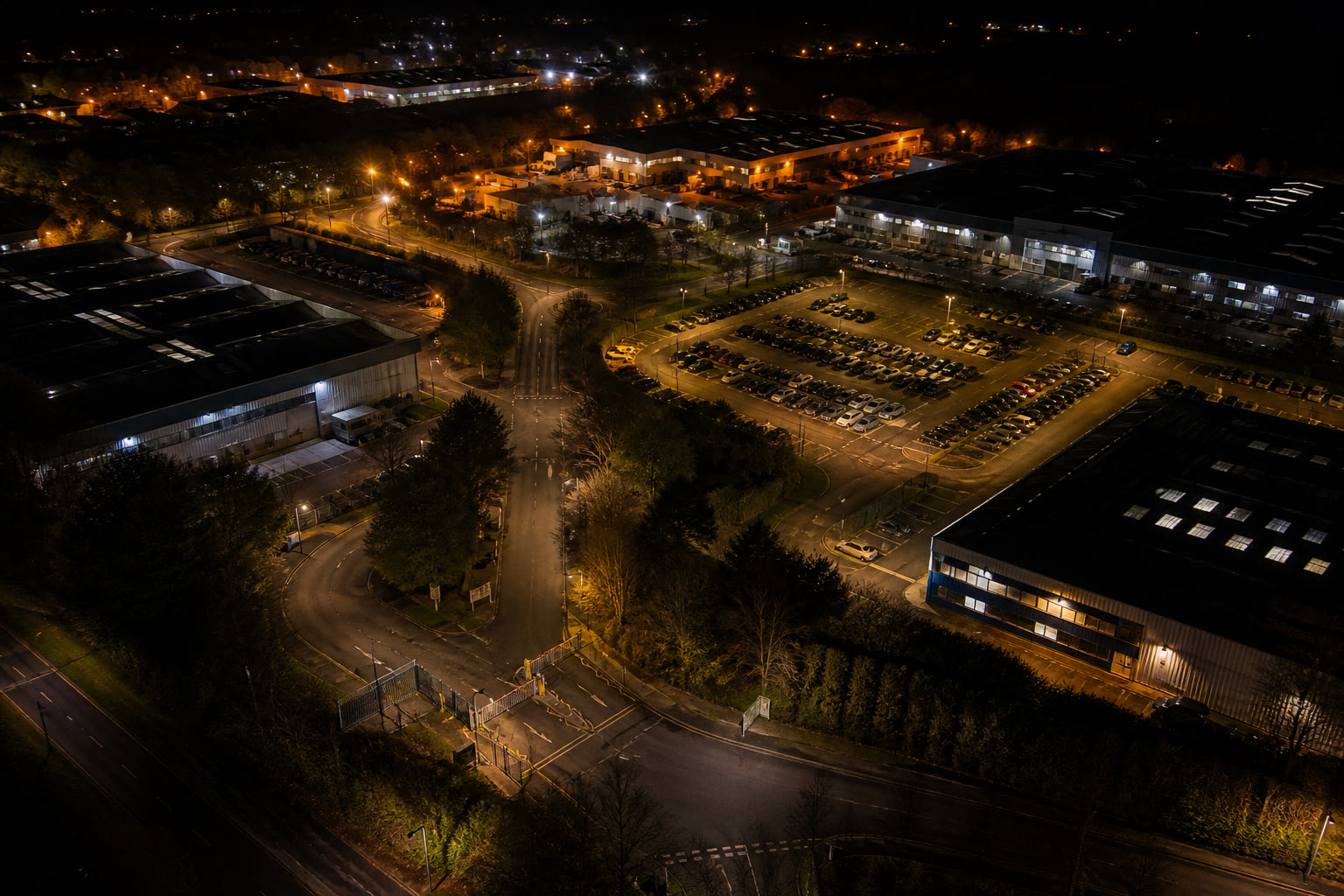 An aerial view of a mixed commercial estate at night, several buildings, internal roads, a barrier-controlled entrance and a lit car park.