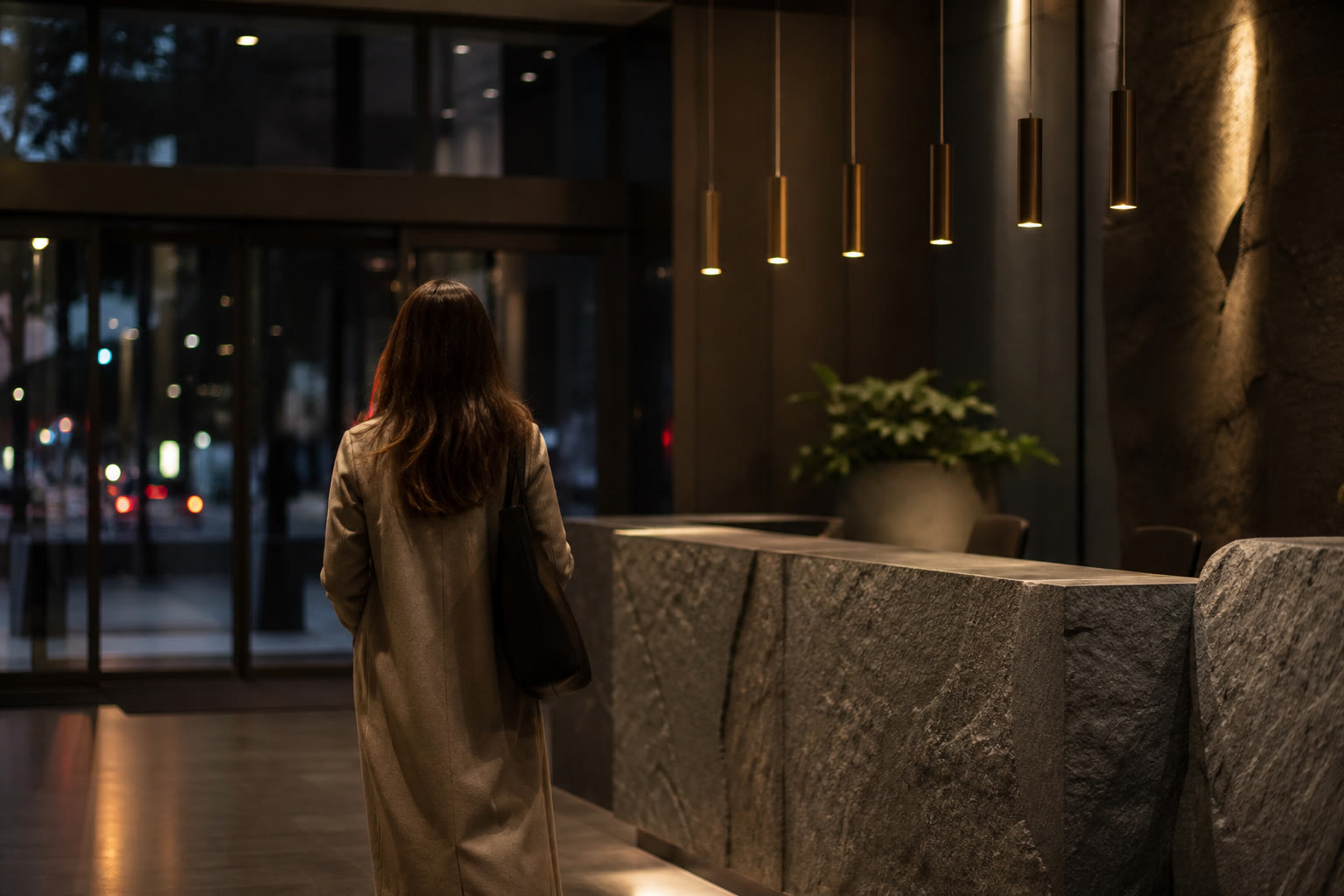 A corporate reception area in the evening, a visitor seen from behind approaching a stone counter under warm pendant lighting, glass entrance doors behind.